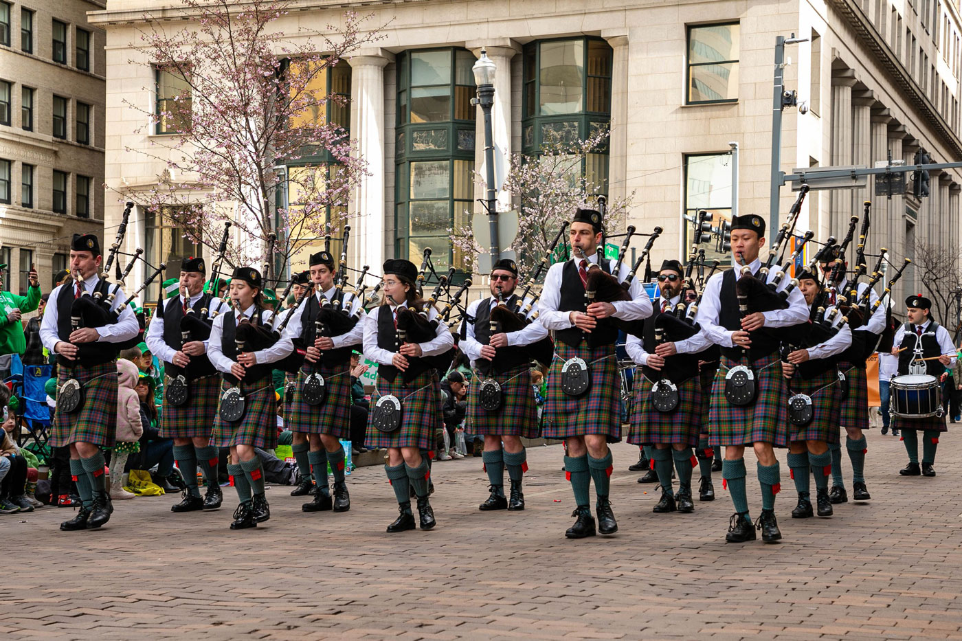 A group of musicians in tartan kilts play bagpipes and march in a city street parade, surrounded by spectators and buildings.